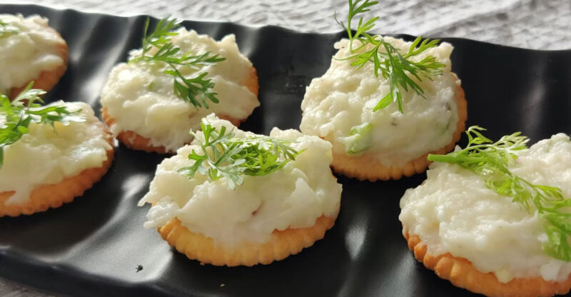 Close-up of Monaco biscuits topped with a creamy mashed potato spread and garnished with fresh coriander leaves, arranged on a black tray