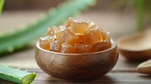 A wooden bowl filled with glossy aloe vera sweet cubes, with fresh aloe leaves in the background.