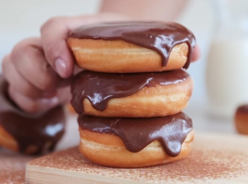 A close-up of three stacked, golden-brown vegan donuts generously coated in a thick, glossy chocolate glaze, with a hand gently picking up the top donut