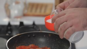 A Person Pouring Milk from A Small Glass Cup Into an Orange Measuring Cup Over a Pan of Cooking Carrot Beetroot Mixture