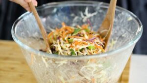 A Pair of Wooden Spoons Tossing Shredded Cabbage, Carrots, and Edamame in A Glass Bowl