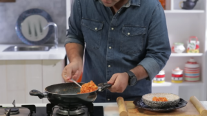 A Man in A Denim Shirt Serving Carrot Beetroot Halwa from A Pan Into a Bowl in A Kitchen