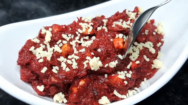 A Plate of Carrot Beetroot Halwa Garnished with Crumbled Nuts and Paneer, with A Spoon Partially Dipped In