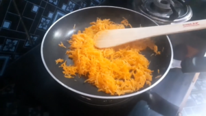 Grated Carrots Being Sautéed in A Black Non-Stick Pan with A Wooden Spatula on A Stovetop