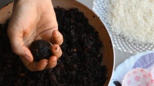 A Hand Holding a Freshly Shaped Carrot Beetroot Laddu Over a Pan of The Mixture