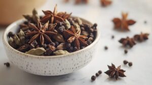 A ceramic bowl filled with whole spices, including star anise, cardamom, black pepper, and cloves, set on a white marble surface