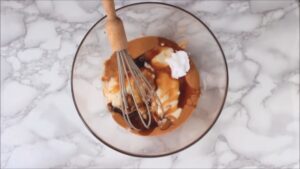A whisk rests in a glass bowl filled with peanut butter, coconut oil, maple syrup, and vanilla extract before mixing