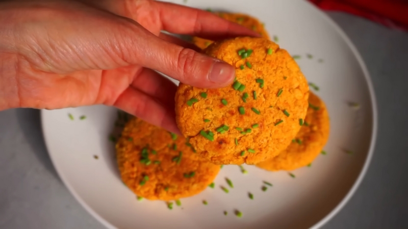 A hand holding a chickpea and sweet potato burger patty topped with chopped herbs over a plate with more patties