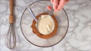 A hand holds a measuring cup of melted coconut oil above a bowl of peanut butter on a marble countertop