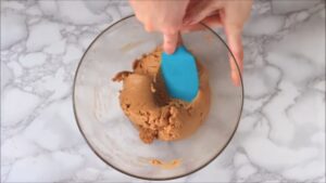 A person uses a blue spatula to mix a thick fudge mixture in a glass bowl on a marble surface