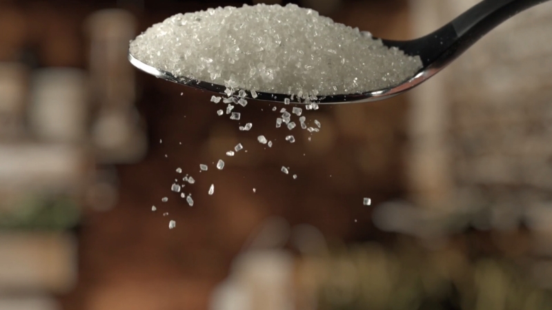 A spoon holding white sugar crystals with some grains falling down