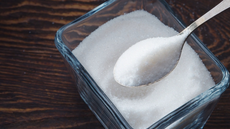 A glass container filled with white sugar and a metal spoon scooping some out