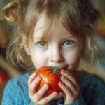 A close-up photo of a young child with light hair holding and eating a large, red tomato