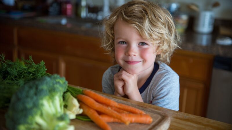 A smiling young boy with curly blonde hair rests his chin on his hands, looking at the camera. In the foreground is a cutting board with fresh broccoli, carrots, and dill