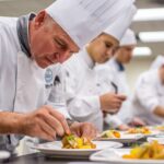 Chefs in white uniforms and tall hats meticulously plating gourmet dishes in a professional kitchen