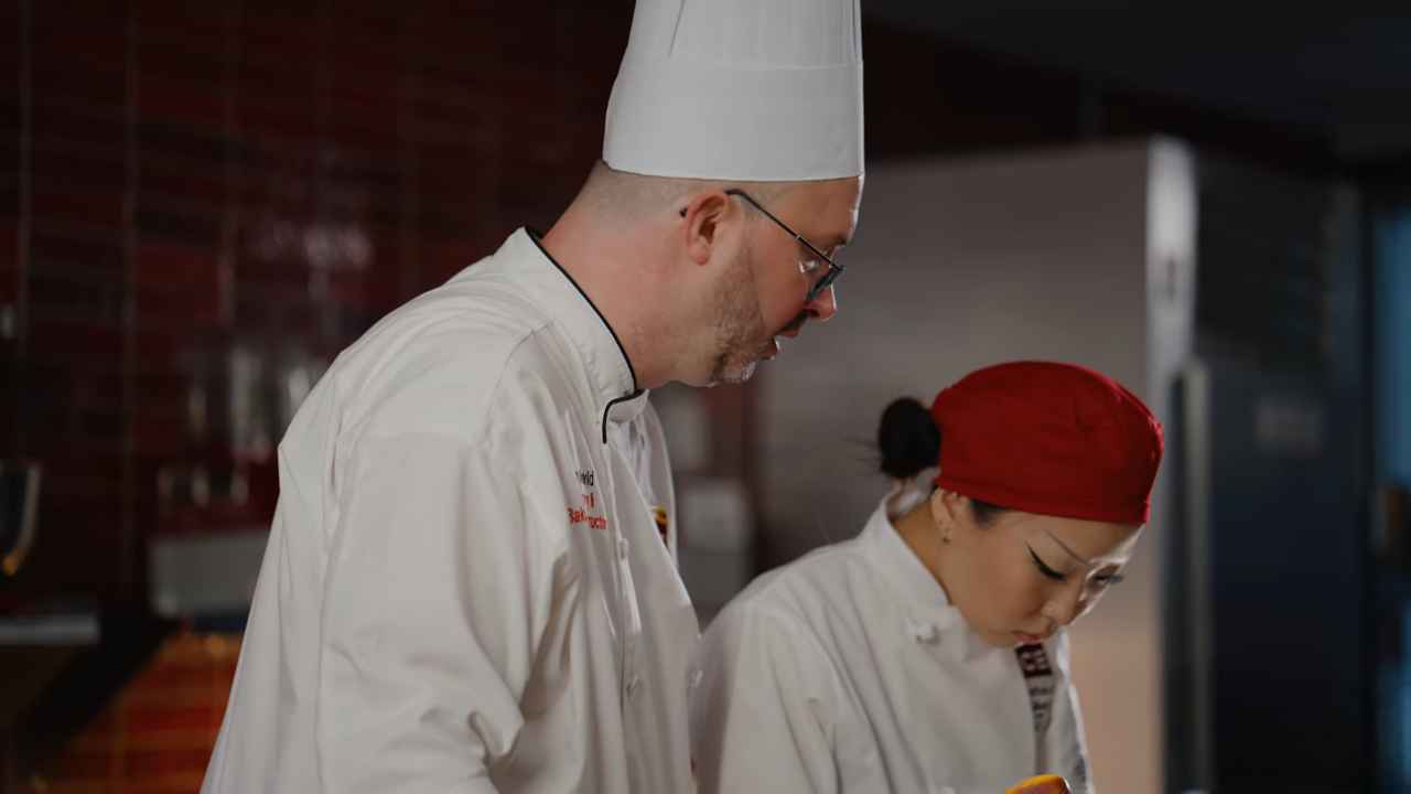Two chefs in professional uniforms working together in a kitchen — one wearing a tall white chef’s hat, the other in a red cap, both focused on food preparation