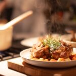 A warm, home-cooked meal with roasted meat and vegetables on a plate, garnished with herbs, beside a pot cooking on the stove