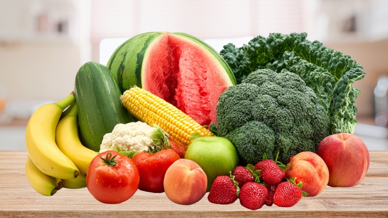 A mix of man-made fruits and vegetables, including bananas, watermelon, corn, tomatoes, broccoli, cauliflower, kale, apples, peaches, and strawberries