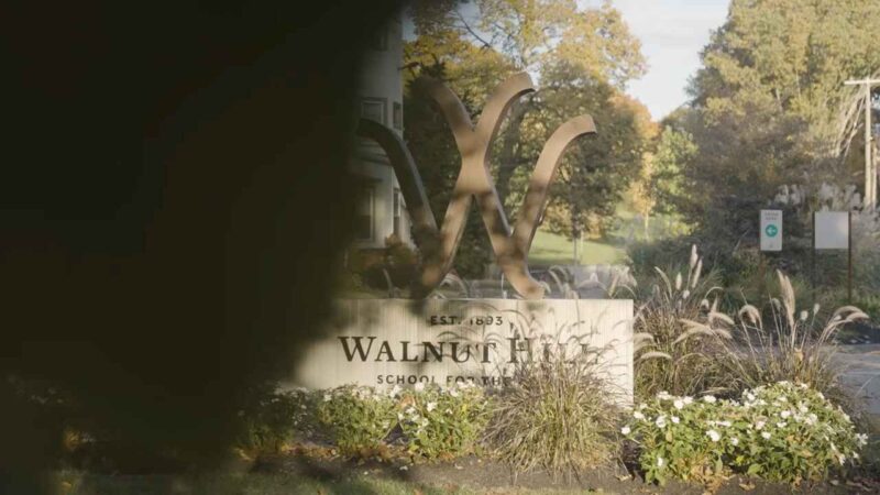 Outdoor sign of Walnut Hill School for the Arts surrounded by greenery and flowers, with a large bronze “W” sculpture and the school’s name engraved on a stone base