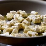 Seasoned tofu cubes sizzling gently in olive oil within a dark non-stick skillet on a stovetop