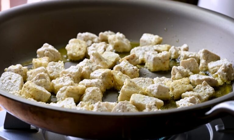 Seasoned tofu cubes sizzling gently in olive oil within a dark non-stick skillet on a stovetop