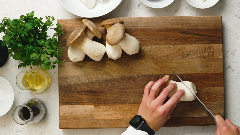 Hand slice king oyster mushroom stems lengthwise on a wooden cutting board before cooking