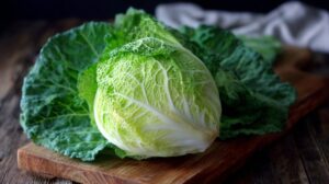 Whole green cabbage resting on a wooden board with loose leaves around it