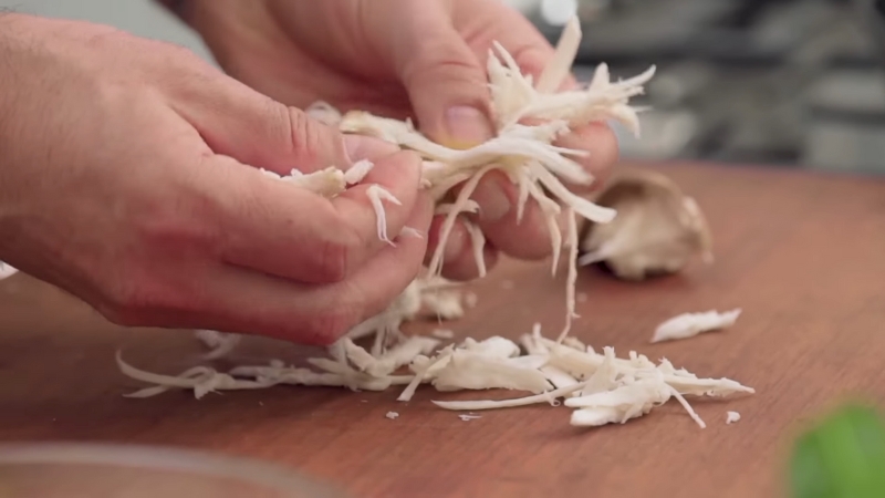 Hands pull cooked king oyster mushroom stems into thin strands on a cutting board