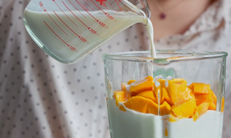 A measuring cup pours milk into a blender filled with sliced mangoes