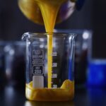 Yellow liquid pours into a clear 250 ml glass beaker on a dark lab table