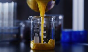 Yellow liquid pours into a clear 250 ml glass beaker on a dark lab table