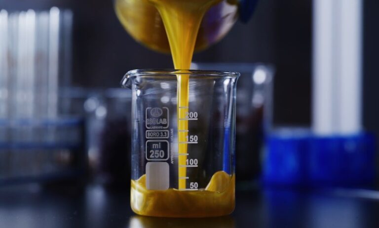 Yellow liquid pours into a clear 250 ml glass beaker on a dark lab table