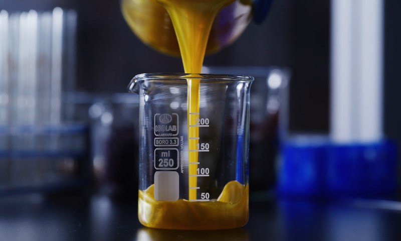 Yellow liquid pours into a clear 250 ml glass beaker on a dark lab table