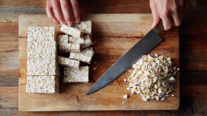 Tempeh being cut into pieces on a wooden cutting board with a knife
