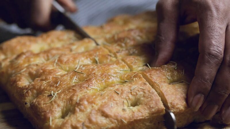 Hands slicing freshly baked rosemary focaccia with a golden, crispy crust.
